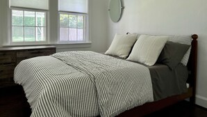 Un dormitorio con cama, dos ventanas con persianas, un espejo redondo en la pared y cabecero de madera.