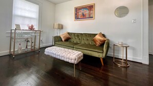 Una sala de estar con un sofá verde, un reposapiés blanco, una mesa de centro de vidrio y una mesita auxiliar. Hay un cuadro en la pared y una ventana con persianas.