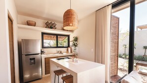 A modern kitchen with a stainless steel refrigerator, a large island with stools, a hanging wicker light, and a view of the garden through large windows. - Ola de Luna, Charming bungalow in El Pescadero with AC, Pool and yoga deck. (El Pescadero)
