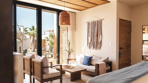 A modern living room with a sofa, chairs, a coffee table, and a large window offering a view of palm trees and a building. - Ola de Luna, Charming bungalow in El Pescadero with AC, Pool and yoga deck. (El Pescadero)