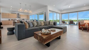 Ein geräumiger Wohnbereich mit grauem Ecksofa, einem Holztisch und großen Fenstern mit Blick ins Freie.
