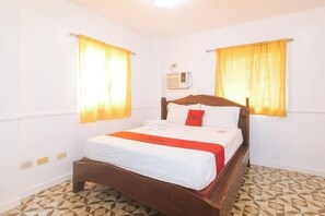 Una cama individual con estructura de madera, ropa de cama blanca y una almohada decorativa roja. Hay un equipo de aire acondicionado instalado en la pared sobre la cama. El cuarto tiene un piso con estampado geométrico y dos ventanas con cortinas amarillas.