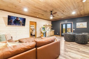 Ein gemütliches Wohnzimmer mit einem braunen Ledersofa, einem grauen Hocker und einer Holzbalkendecke mit Deckenventilator.