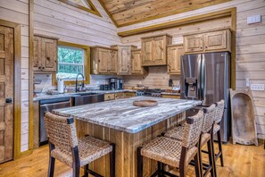 A kitchen with wooden cabinets, a large island with bar stools, a stainless steel refrigerator, and a window with a view of trees. - New Luxury Cabin in Coosawattee River Resort _ Hot Tub + Views + Sleeps 12 (Ellijay)