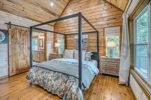 A four-poster bed with a floral bedspread, wooden walls, and a large window with a view of trees. - New Luxury Cabin in Coosawattee River Resort _ Hot Tub + Views + Sleeps 12 (Ellijay)