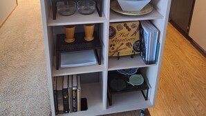Une étagère blanche contenant divers ustensiles de cuisine, notamment des verres à vin, une machine à café et un livre de recettes intitulé « Apéritifs rapides ».