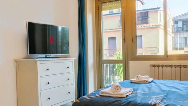 A hotel room with a bed, a TV on a white dresser, a window with a view of buildings, and a radiator.