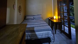 A bedroom with a wooden ceiling, a bed with patterned bedding, a bedside table with a lamp, and a window with a view of greenery.