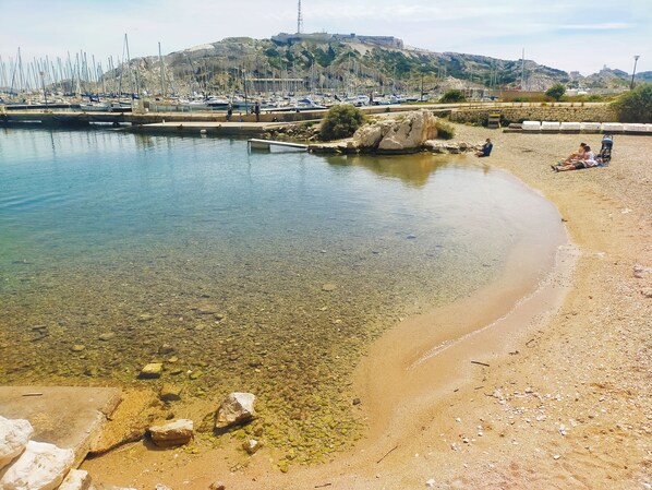 A beach with clear shallow waters, rocky shore, and a distant view of boats and a hill. - Studio on Frioul Island: at the foot of a beach and the Morgiret calanque. (Marseille)