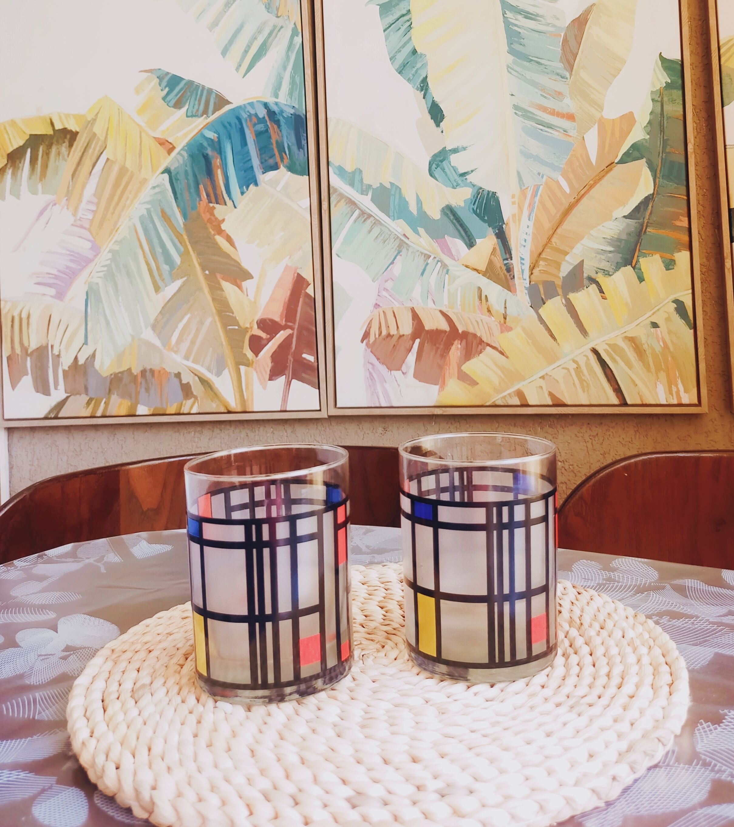 A table with two decorative glass candle holders on a woven placemat, set against a wall with abstract paintings.