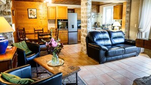 Una sala de estar acogedora con un sofá de cuero negro, una mesa de comedor de madera y sillas. Se ve una cocina con armarios de madera y un refrigerador al fondo.