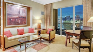 Habitación de hotel con sofá, sillones, una mesa de centro y una mesa de comedor. Se observa un cuadro grande en la pared y una vista de la ciudad a través de la ventana.