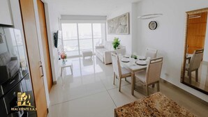 A modern living room with a sofa, dining table, and chairs. There is a TV mounted on the wall, a clock, and a window with a view of the city. - Luxurious & Modern Apartment 1503 Building h2 (Cartagena de Indias)