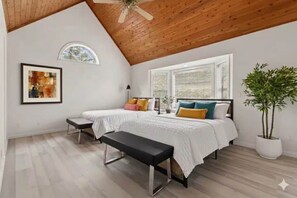Une chambre avec un plafond en pente, un lit agrémenté de plusieurs coussins, un banc, une plante en pot et un tableau encadré au mur.