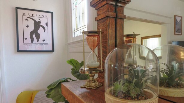 Une pièce avec une table en bois, une œuvre d’art encadrée, une fenêtre et une plante placée sous une cloche de verre.