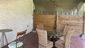 A covered outdoor seating area with wicker chairs and a glass table, brick walls, and a wooden partition.