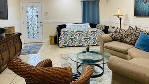 A living room with a sofa, two wicker chairs, a glass coffee table, a patterned bedspread, a door with a decorative glass panel, and a blue curtain.