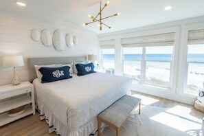 A bedroom with a large bed, a bench, a nightstand, a lamp, and a view of the beach.