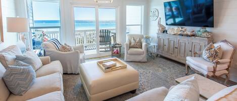 A spacious living room with a large TV, comfortable seating, a view to the ocean, and a balcony.