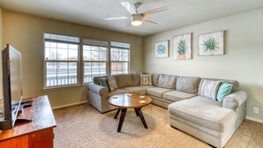 A living room with a sectional sofa, a coffee table, and a ceiling fan. There are three windows and a TV on a stand.