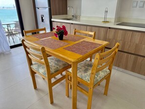 A wooden dining table with four chairs, a small potted plant, and a kitchen in the background. - Sunset hills Apartments (Mazatlán)