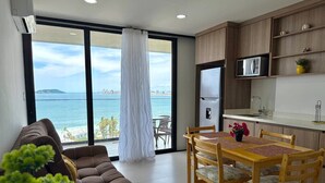 A modern living room with a sofa, dining table, and chairs, a kitchen area with a microwave, and a large window with a view of the sea. - Sunset hills Apartments  (Mazatlán)