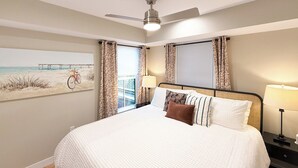 Une chambre à coucher avec un lit, un ventilateur de plafond, une table de chevet avec une lampe et un tableau représentant une scène de plage.