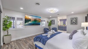 A bedroom with a large bed, a painting of a landscape, a potted plant, and a bathroom visible in the background.