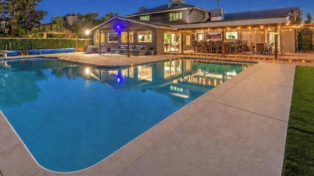 A swimming pool with a patio area, a covered seating area, and a house in the background.