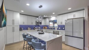 A modern kitchen with white cabinets, a central island with bar stools, a stainless steel refrigerator, and a blue-tiled backsplash.