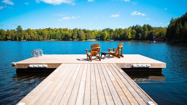 Une jetée en bois, agrémentée de deux fauteuils Adirondack et d’une table, offre une vue sur un lac paisible dont les rives sont bordées d’une forêt.