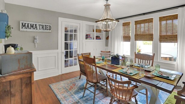 A dining area with a wooden table set for four, surrounded by chairs. A chandelier hangs above, and there are large windows with bamboo blinds. A sign on the wall reads "MAMA TRIED."