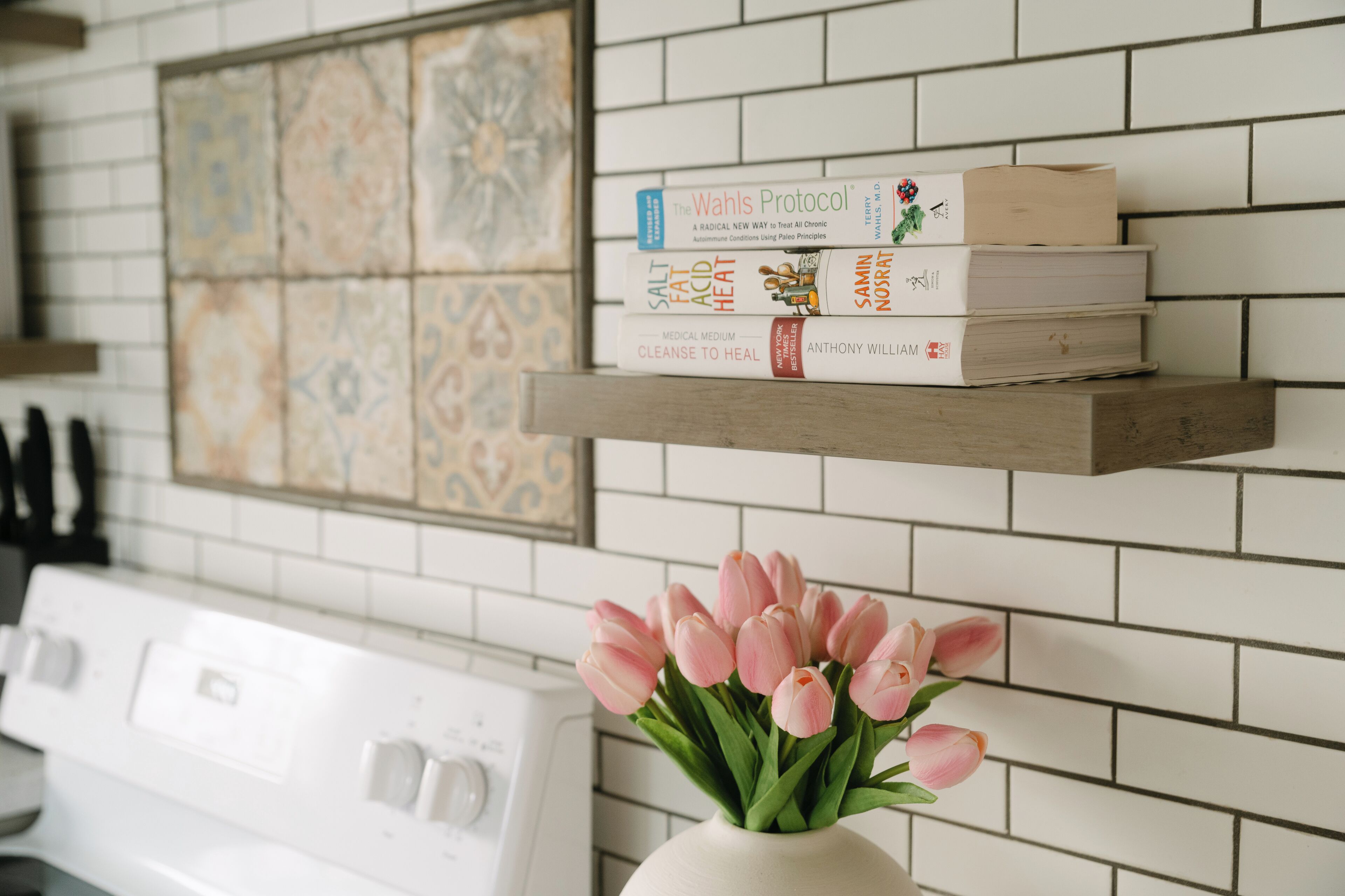 Una cocina con estufa blanca, un estante con libros y un florero con tulipanes rosados.