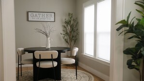 A dining area with a round table, four chairs, a vase with branches, a framed sign, and a window with a plant. - Luxe Cottage, Near Downtown and Ocean, Backyard Fire Pit, King Bed (Corpus Christi)