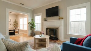 A living room with a fireplace, a television mounted above it, and a dining area in the background. - Luxe Cottage, Near Downtown and Ocean, Backyard Fire Pit, King Bed (Corpus Christi)