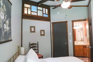 Une chambre à coucher comprenant un lit, une chaise, un ventilateur de plafond et une porte donnant sur une salle de bain.