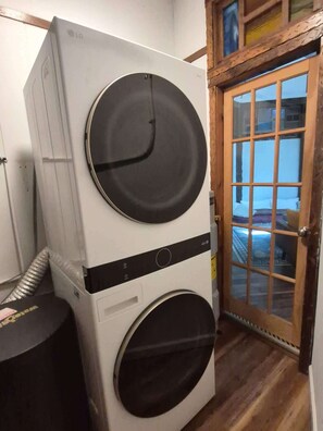 Une machine à laver et un sèche-linge superposés dans une pièce dotée d’une porte en bois et offrant une vue sur un bâtiment à l’extérieur.