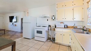 A compact kitchen with white appliances, a small dining table, and a TV mounted on the wall.