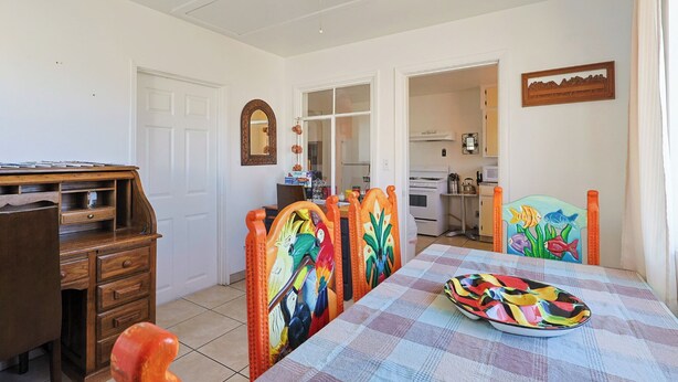 A dining area with a checkered tablecloth, wooden chairs with colorful fish designs, a wooden desk, and a kitchen in the background.