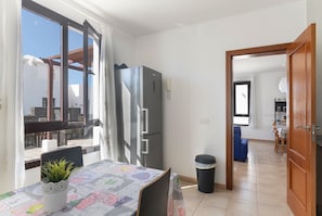 A dining area with a table, chairs, a refrigerator, a trash bin, and a view through a window.