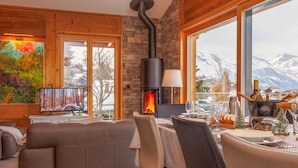 Ett mysigt vardagsrum med en öppen spis, en soffa, ett matbord för två och ett stort fönster med utsikt över bergen.