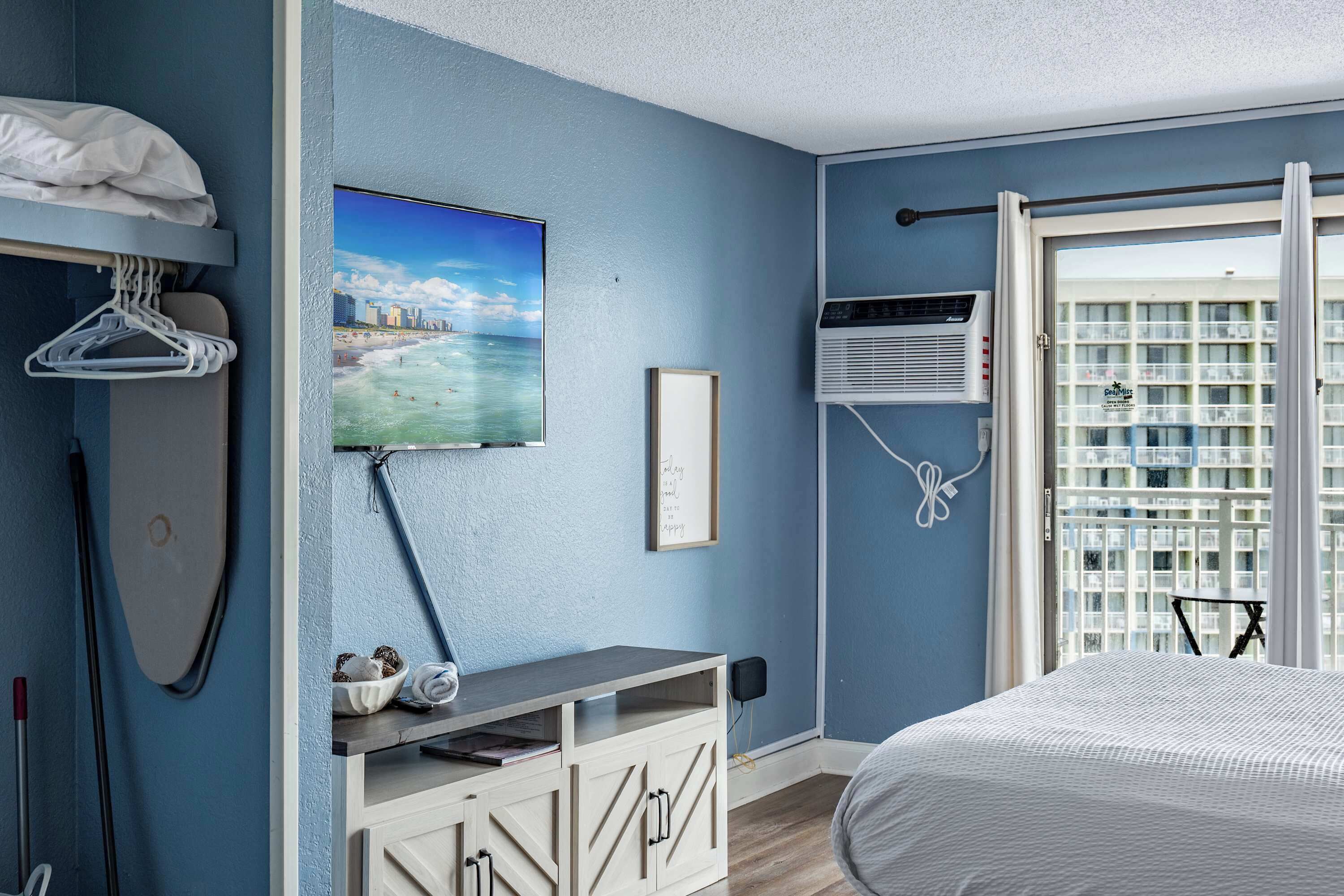 A bedroom with a bed, a painting of a beach scene, a wall-mounted air conditioner, and a view of a building through a window.