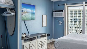 A bedroom with a bed, a painting of a beach scene, a wall-mounted air conditioner, and a view of a building through a window. - Ocean View Condo Sleeps 4! (Myrtle Beach)