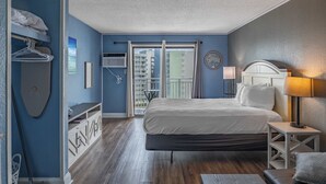 A hotel room with a bed, a bedside table, a lamp, a window with a view of buildings, and a door leading to a balcony. - Ocean View Condo Sleeps 4! (Myrtle Beach)