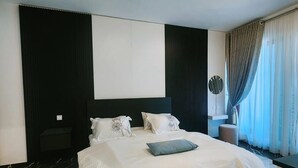 Une chambre d’hôtel moderne avec un grand lit, une table de chevet, une commode, une fenêtre avec des rideaux et une œuvre d’art accrochée au mur.