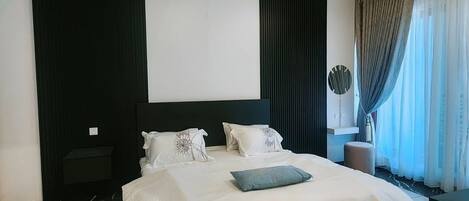 Ein modernes Hotelzimmer mit einem großen Bett, einem Nachttisch, Vorhängen vor einem Fenster und einem an der Wand befestigten Kunstwerk.