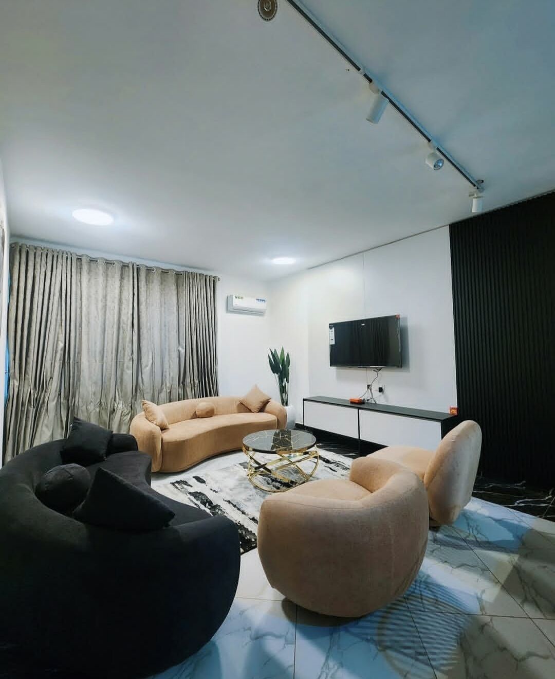 A modern living room with a sofa, armchair, and bean bag, a flat-screen TV mounted on the wall, and a large window with curtains.