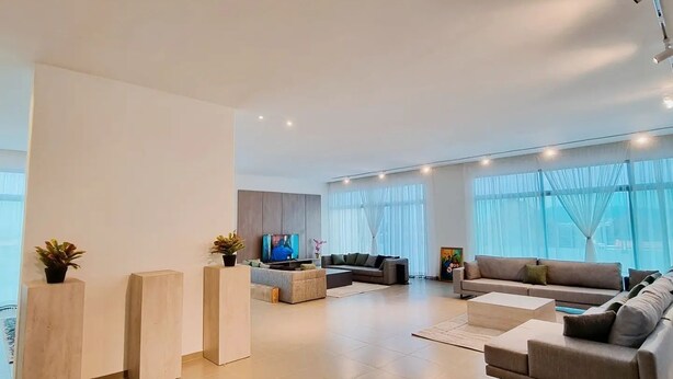 A spacious living room with a large sofa set, a coffee table, and a TV. There are potted plants and a modern light fixture on the ceiling. - Boya Apartments (Abuja)