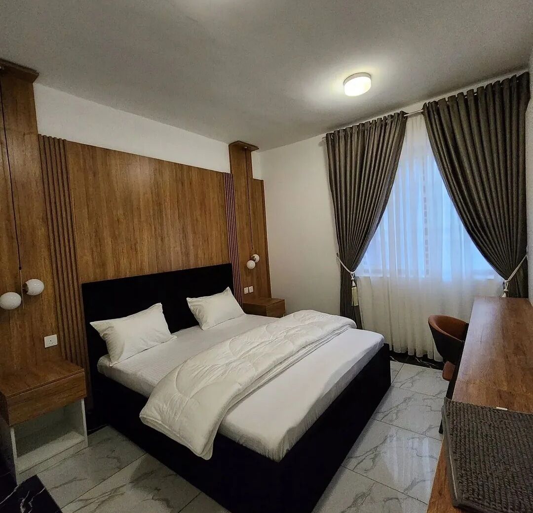 A modern hotel room with a large bed, wooden headboard, two bedside tables, a desk, and a chair.