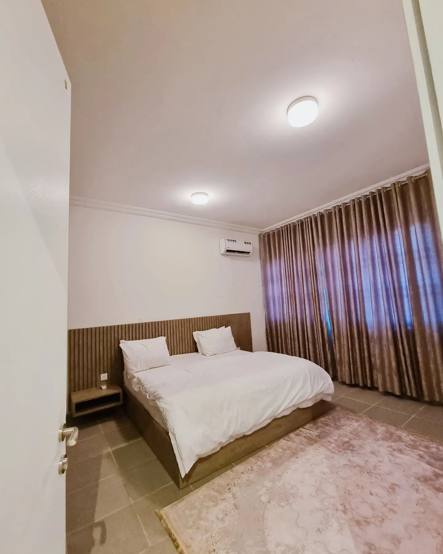 A hotel room with a bed, a nightstand, an air conditioning unit, a window with curtains, and a tiled floor.